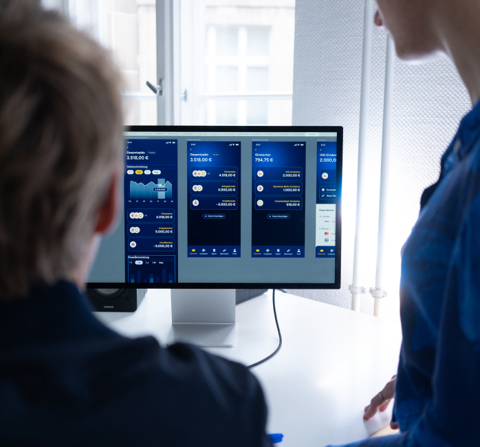 Two designner from Ennostudio designers reviewing mobile app interface screens with charts and balance data on a desktop monitor.