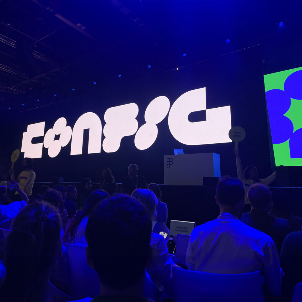 A large audience sitting in front of a stage with a big screen. The screen behind shows the titel of the event "Config".
