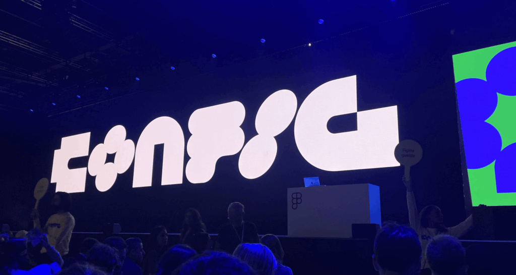 A large audience sitting in front of a stage with a big screen. The screen behind shows the titel of the event "Config".